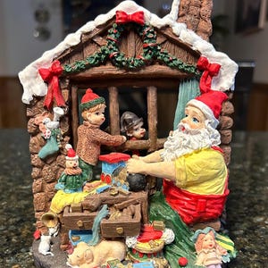 May include: A detailed Christmas figurine depicting Santa Claus in a workshop setting. The figurine features a brick-like exterior, a snow-covered roof, and a window with children. Santa is wearing a red hat and yellow shirt, surrounded by toys and gifts.