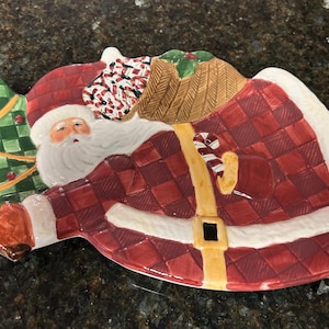 May include: A ceramic Santa Claus-shaped serving platter. The platter features a red and white plaid design on Santa's coat, a white beard, and a red hat. A green Christmas tree is on the left side of the platter.