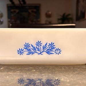 May include: A rectangular, white casserole dish with a blue floral design. The dish has a slightly flared shape and small handles on each end. The blue floral pattern is centered on the front of the dish.