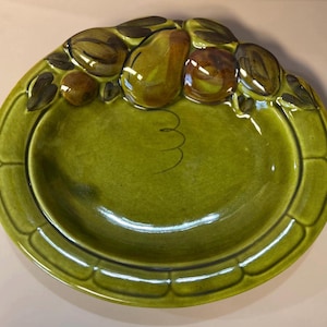 May include: A green ceramic serving dish with a raised fruit and leaf design around the rim. The dish features a glossy finish and a decorative swirl in the center. The fruit includes pears and other fruits.