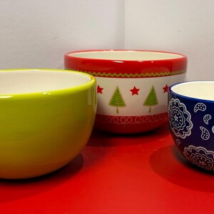 May include: Three ceramic bowls in different sizes and colors. The largest bowl is red with a white band and a Christmas tree design. The medium bowl is bright yellow. The smallest bowl is blue with a white paisley design.