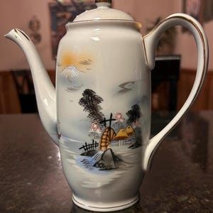 May include: White ceramic coffee pot with a gold-colored handle and spout trim. The pot features a hand-painted landscape scene with a mountain, trees, and a watermill. The lid is topped with a gold-colored knob.