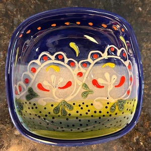 May include: A square, ceramic bowl with a deep blue exterior and a decorative interior. The interior features a white design with red, yellow, and green accents. The bowl is ideal for serving snacks or small dishes.