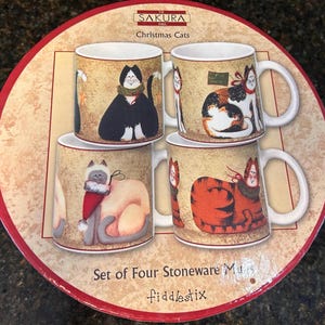 May include: Set of four stoneware mugs with cat illustrations. Each mug features a different cat design, including a black cat, a calico cat, a gray cat, and a red tabby cat. The mugs are on a round, red-edged cardboard base.