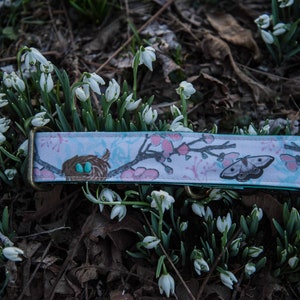 May include: A light blue and white patterned dog collar with a bird's nest, a butterfly, and pink flowers. The collar is lying on a bed of white snowdrop flowers.