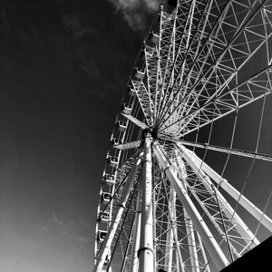 Könnte beinhalten: Ein Schwarzweißfoto eines großen Riesenrads vor einem bewölkten Himmel. Das Rad besteht aus Metall und hat viele Speichen. Das Rad ist teilweise von einem dunklen Objekt im Vordergrund verdeckt.