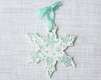 May include: A white snowflake ornament with a teal snowflake design and a teal ribbon for hanging. The ornament is made of ceramic or a similar material and is a winter holiday decoration.