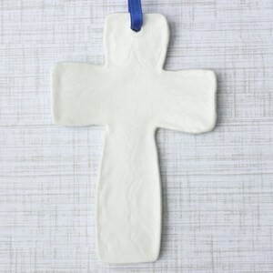 May include: A white ceramic cross with a blue ribbon for hanging. The cross has a simple, minimalist design and is likely intended for religious or decorative purposes. The cross is approximately 6 inches long.