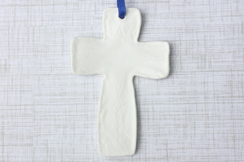 May include: A white ceramic cross with a blue ribbon for hanging. The cross has a simple, minimalist design and is likely intended for religious or decorative purposes. The cross is approximately 6 inches long.