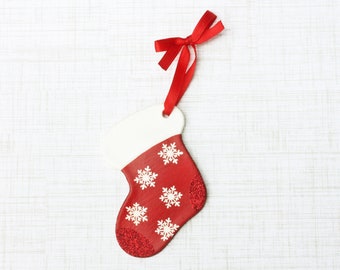 May include: A red Christmas stocking ornament with white snowflake designs and a white cuff. The stocking is adorned with red glitter at the bottom and has a red ribbon for hanging. Festive holiday decor.