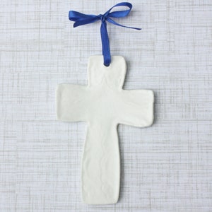 May include: A white ceramic cross ornament with a blue satin ribbon for hanging. The cross has a simple, minimalist design and is displayed against a light, textured background. The ribbon is tied in a bow at the top.