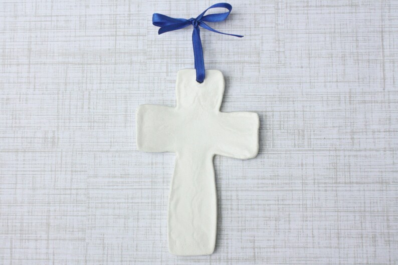 May include: A white ceramic cross ornament with a blue satin ribbon for hanging. The cross has a simple, minimalist design and is displayed against a light, textured background. The ribbon is tied in a bow at the top.