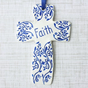 May include: A white ceramic cross with blue floral designs and the word "Faith" in blue script. A blue ribbon is attached for hanging. The cross is a religious decorative item.