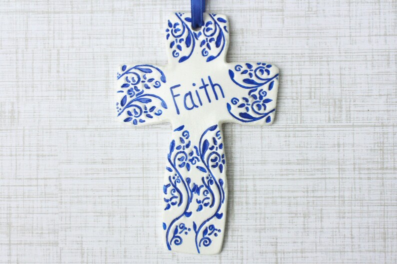 May include: A white ceramic cross with blue floral designs and the word "Faith" in blue script. A blue ribbon is attached for hanging. The cross is a religious decorative item.