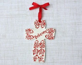 May include: A white ceramic cross ornament with red floral designs and the word "Believe". It has a red ribbon for hanging. The ornament is a religious item.