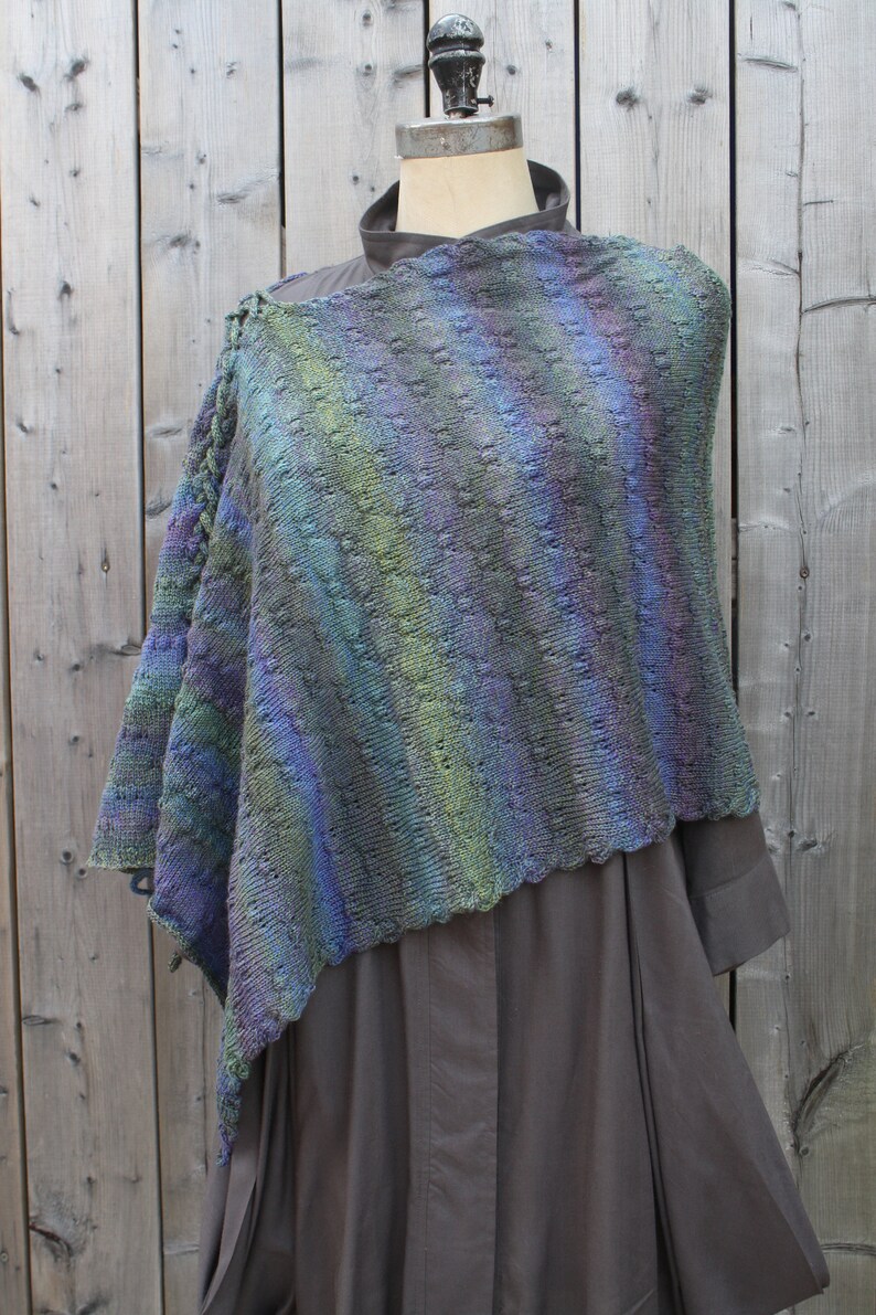 May include: A blue, green, and purple knitted shawl with a lacy pattern. The shawl is draped over a mannequin wearing a dark gray dress.