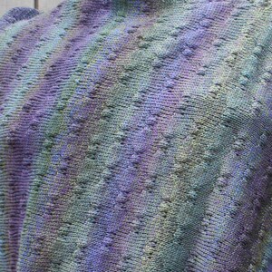 May include: A close-up of a knitted shawl with a purple, green, and blue color gradient. The shawl has a lacy pattern and is made with a soft yarn.