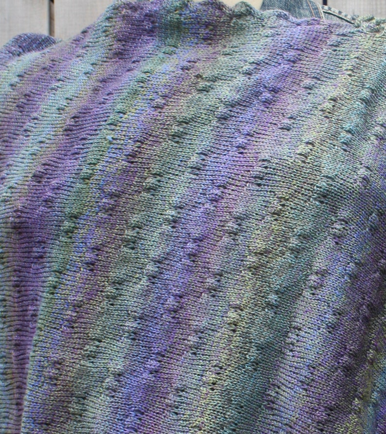 May include: A close-up of a knitted shawl with a purple, green, and blue color gradient. The shawl has a lacy pattern and is made with a soft yarn.