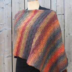 May include: A knitted poncho with a striped pattern in shades of orange, red, brown, and gray. The poncho is draped over a mannequin and has a triangular shape.