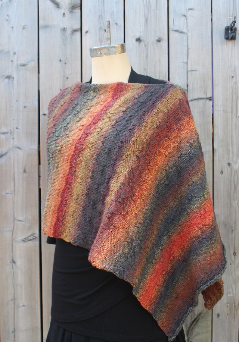May include: A knitted poncho with a striped pattern in shades of orange, red, brown, and gray. The poncho is draped over a mannequin and has a triangular shape.