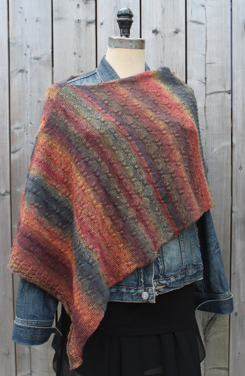 May include: A striped knit shawl in shades of red, orange, brown, and gray. The shawl is draped over a mannequin wearing a denim jacket and a black skirt.