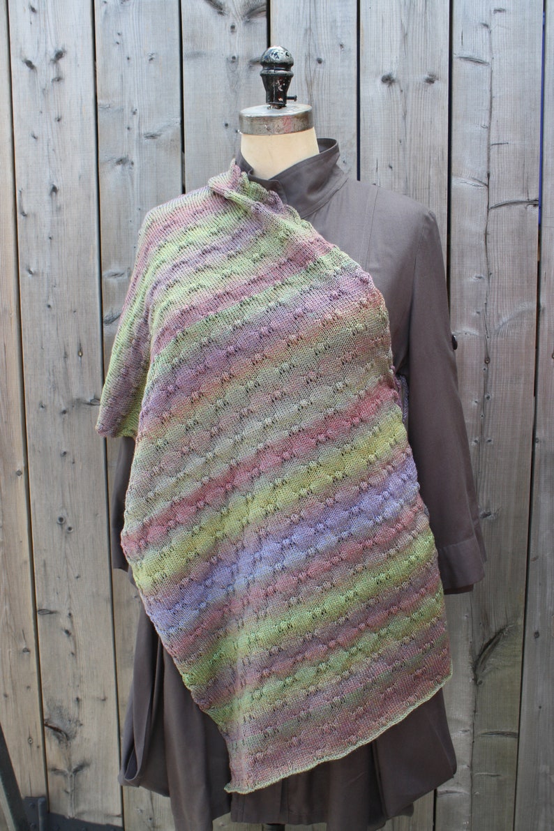May include: A multi-colored knitted shawl with a textured pattern. The shawl is draped over a mannequin wearing a brown shirt.