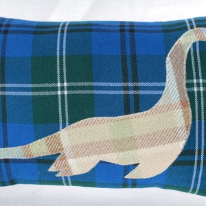 May include: A rectangular pillow with a blue, green, and white plaid pattern. A beige and brown Nessie-like creature is appliquéd on the front. The pillow is a decorative item, perfect for a couch or bed.