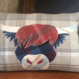 May include: A decorative pillow with a Highland Cow design. The pillow is made of a beige and white plaid fabric with a red, blue, and white plaid cow face appliqué. The pillow has a white tag with the text "The Thistle Sea Navy" and a graphic of a thistle.