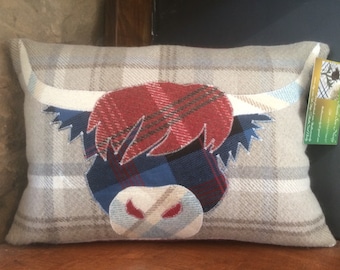 May include: A decorative pillow with a Highland Cow design. The pillow is made of a beige and white plaid fabric with a red, blue, and white plaid cow face appliqué. The pillow has a white tag with the text "The Thistle Sea Navy" and a graphic of a thistle.