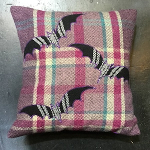 May include: Purple and white plaid pillow with three black bats embroidered on it. The bats have white stripes and purple outlines.