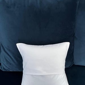 May include: A white pillow with a folded edge rests in front of two dark blue velvet pillows. The pillows are a deep shade of blue, creating a contrast with the white pillow.