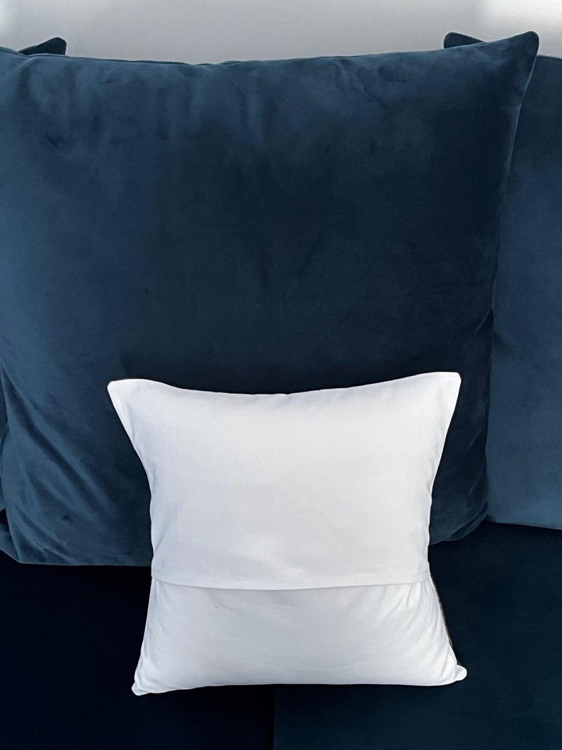 May include: A white pillow with a folded edge rests in front of two dark blue velvet pillows. The pillows are a deep shade of blue, creating a contrast with the white pillow.