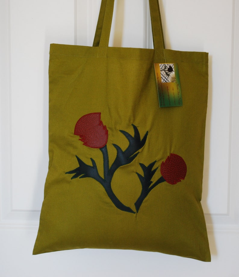 May include: A green canvas tote bag with a black and red leather applique of a stylised flower. The bag has two long straps.