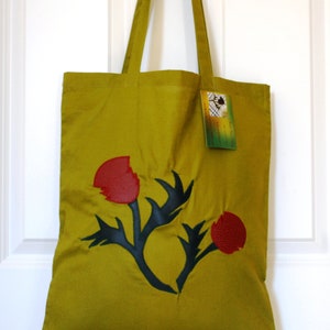 May include: A green tote bag featuring a black and red floral design. The bag has a tag that reads "Handmade in New Zealand".