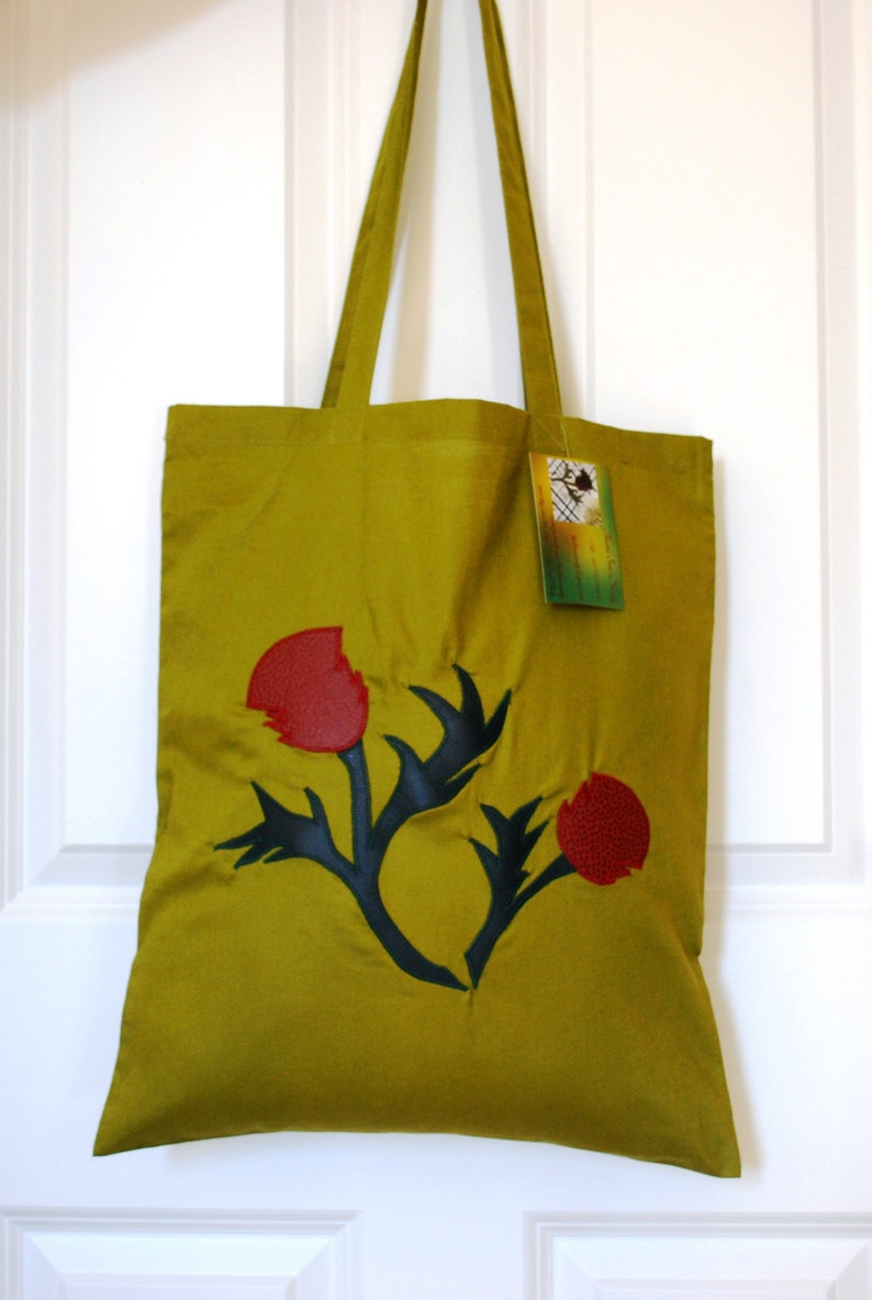 May include: A green tote bag featuring a black and red floral design. The bag has a tag that reads "Handmade in New Zealand".