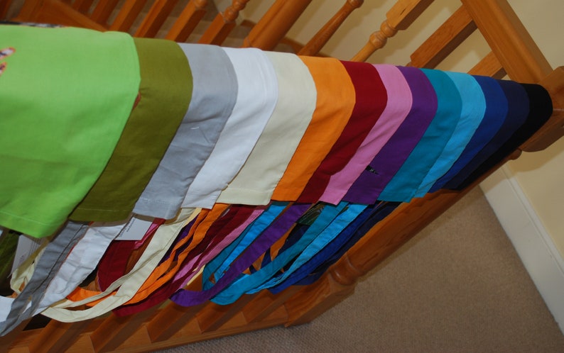 May include: A collection of fabric scraps in various colours, including green, olive, grey, white, cream, orange, red, pink, purple, blue, teal, and black. The fabric scraps are arranged in a rainbow pattern and are hanging from a wooden railing.