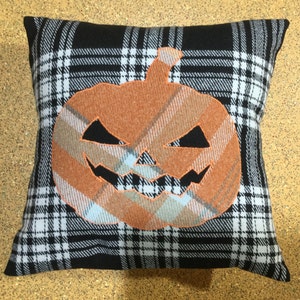 May include: A black and white plaid pillow with a large orange pumpkin applique. The pumpkin has a carved jack-o'-lantern face.