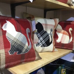 May include: Plaid throw pillows with cat silhouettes. The pillows feature a red, cream, and brown plaid pattern. Each pillow has a cat silhouette in white, black, or cream. The pillows are displayed on a wooden shelf.