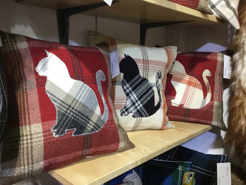 May include: Plaid throw pillows with cat silhouettes. The pillows feature a red, cream, and brown plaid pattern. Each pillow has a cat silhouette in white, black, or cream. The pillows are displayed on a wooden shelf.