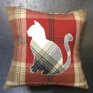 May include: Square decorative pillow with a red and brown plaid pattern. A white cat silhouette is centered on the pillow, with a plaid pattern within the cat shape. The pillow is a cozy home decor item.