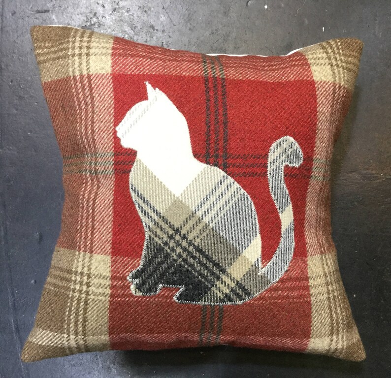 May include: Square decorative pillow with a red and brown plaid pattern. A white cat silhouette is centered on the pillow, with a plaid pattern within the cat shape. The pillow is a cozy home decor item.