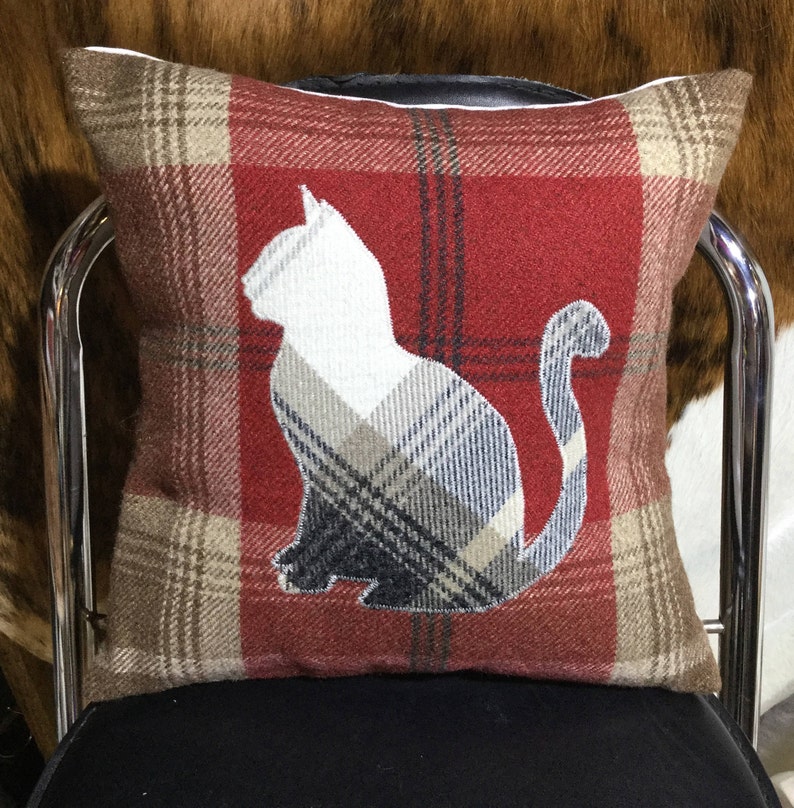 May include: Square decorative pillow with a red plaid pattern and a white cat silhouette. The cat's body is filled with a plaid pattern of beige, gray, and navy blue. The pillow is on a black chair.