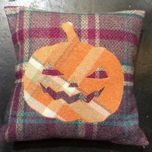 May include: A square pillow with a plaid pattern in shades of burgundy, teal, and beige. A large orange pumpkin with a carved jack-o'-lantern face is appliqued on the front of the pillow.
