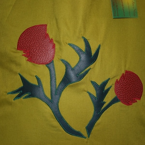 May include: A green fabric background with a dark blue and green embroidered thistle design. The thistle has three red flower buds.