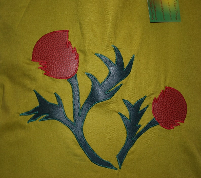 May include: A green fabric background with a dark blue and green embroidered thistle design. The thistle has three red flower buds.