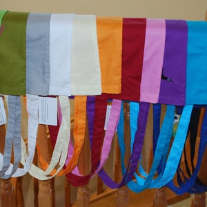 May include: A collection of 14 colourful tote bags with different coloured straps hanging on a wooden railing. The bags are made of a soft, fabric material and feature a variety of colours, including green, grey, white, orange, red, pink, purple, blue, and black.