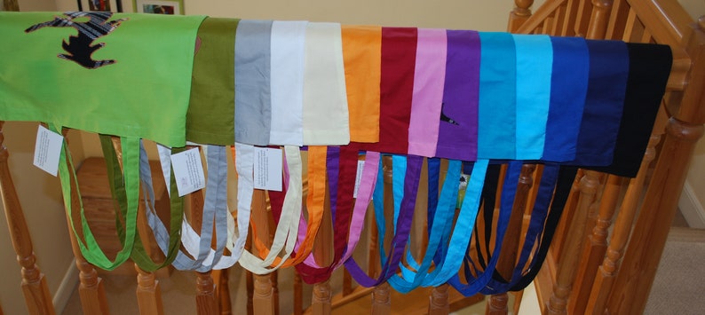 May include: A collection of 14 colourful tote bags with different coloured straps hanging on a wooden railing. The bags are made of a soft, fabric material and feature a variety of colours, including green, grey, white, orange, red, pink, purple, blue, and black.