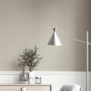 Könnte beinhalten: Ein Raum in neutralen Tönen mit einem strukturierten Tapetenhintergrund. Eine silberne Lampe mit konischem Schirm erstreckt sich von rechts. Ein Holzschrank enthält eine Vase mit Grün, eine Glasvase und ein dekoratives Glas. Der Raum hat eine moderne, minimalistische Ästhetik.