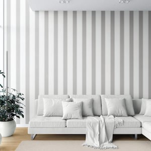 Könnte beinhalten: Ein modernes Wohnzimmer mit einem weißen Ecksofa, Kissen und einer Decke. Die Wand hat vertikale graue und weiße Streifen. Eine Topfpflanze und ein neutraler Teppich vervollständigen die Szene.