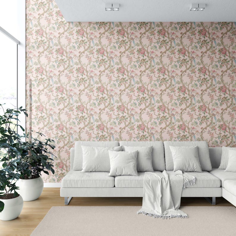 May include: A living room with a white sectional sofa, a light beige rug, and potted plants. The wallpaper features a floral pattern with pink roses and green leaves on a light pink background. The room is well-lit with natural light.